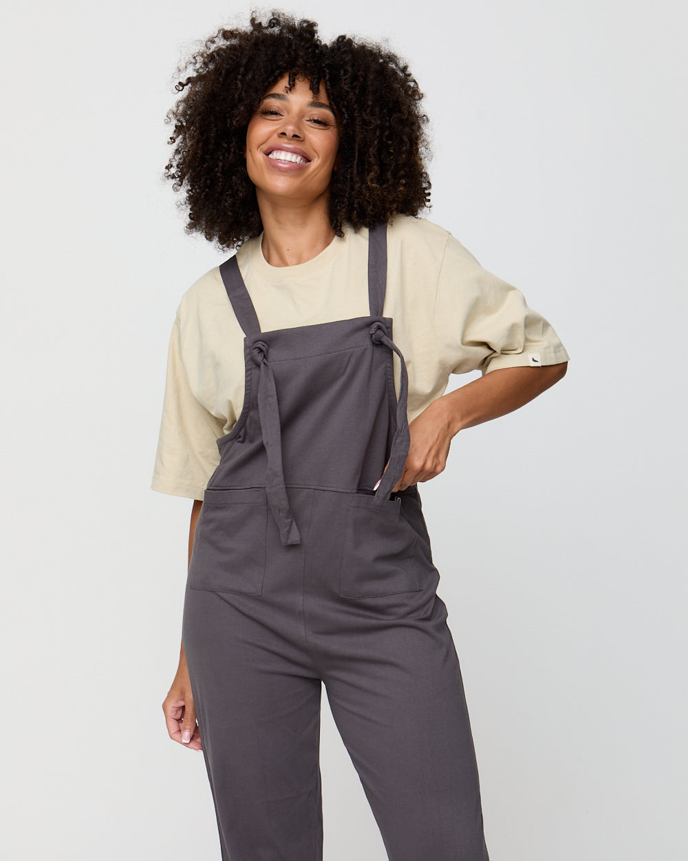 Woman wearing gray overalls and a beige shirt on a plain background