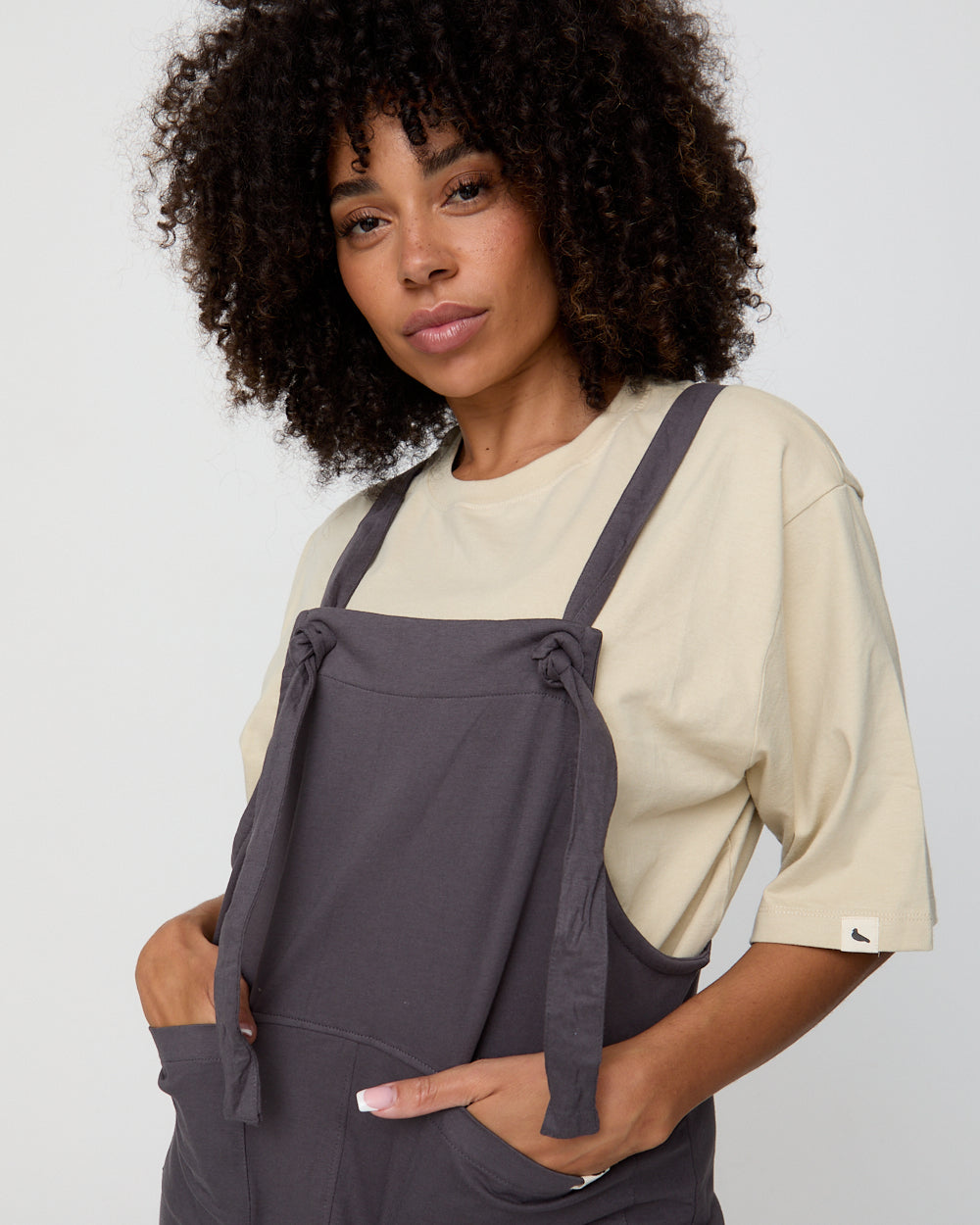 Woman wearing a beige shirt and gray overalls against a white background