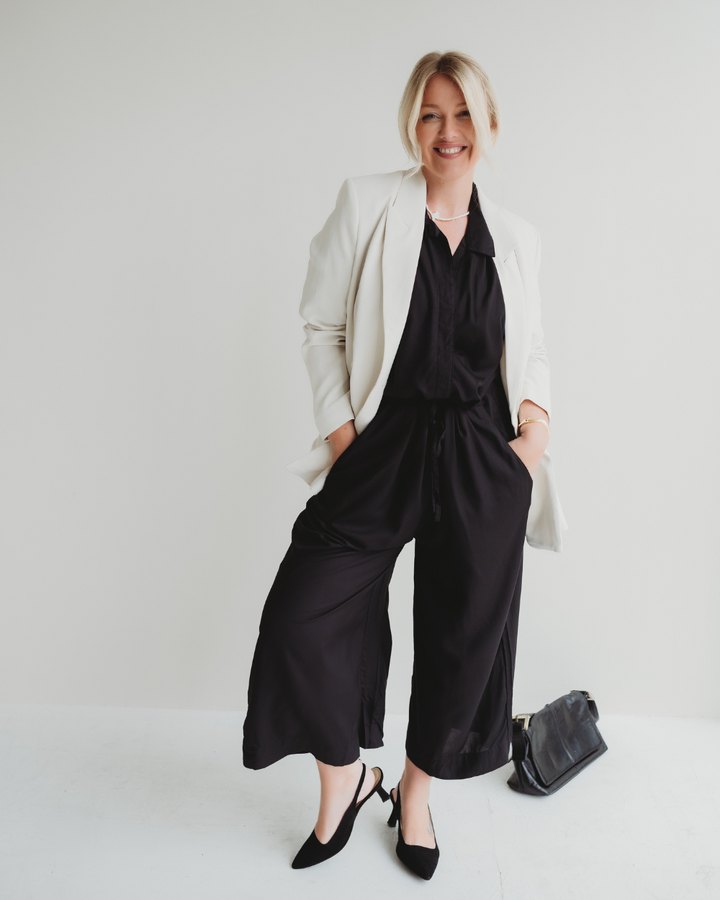 A smiling person stands in a white room, wearing the Iris Black Jumpsuit made of EcoVero viscose with a drawstring waist, under a chic white blazer and black heels. Their hands are in their pockets, and a stylish black bag rests on the floor beside them.