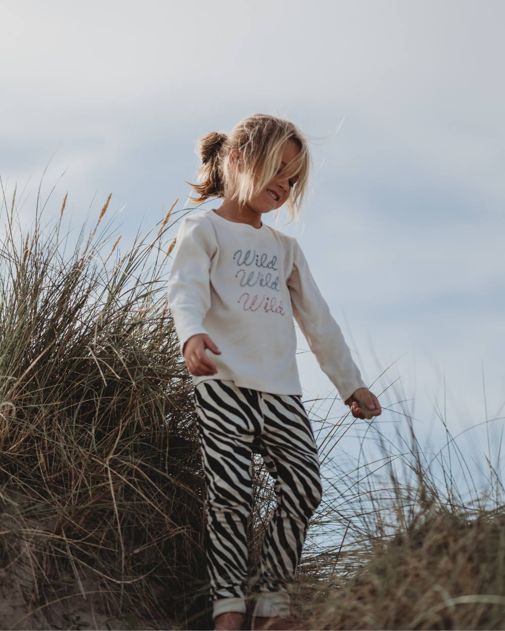 A young blonde girl enjoys a sunny day in tall grass, embracing sustainable fashion with Wild Wild Wild on her white sweatshirt and Zebra Skin leggings made from organic cotton. The sky is mostly clear, dotted with a few clouds.