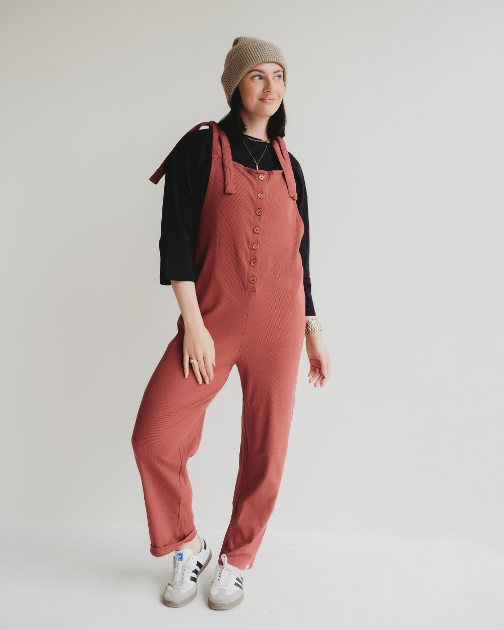 A woman in a casual outfit poses stylishly against a white background wearing Erin Brick Dungarees over a black shirt. She completes her look with a beige knit beanie and white sneakers, embodying laid-back charm and sustainability through the choice of organic cotton.