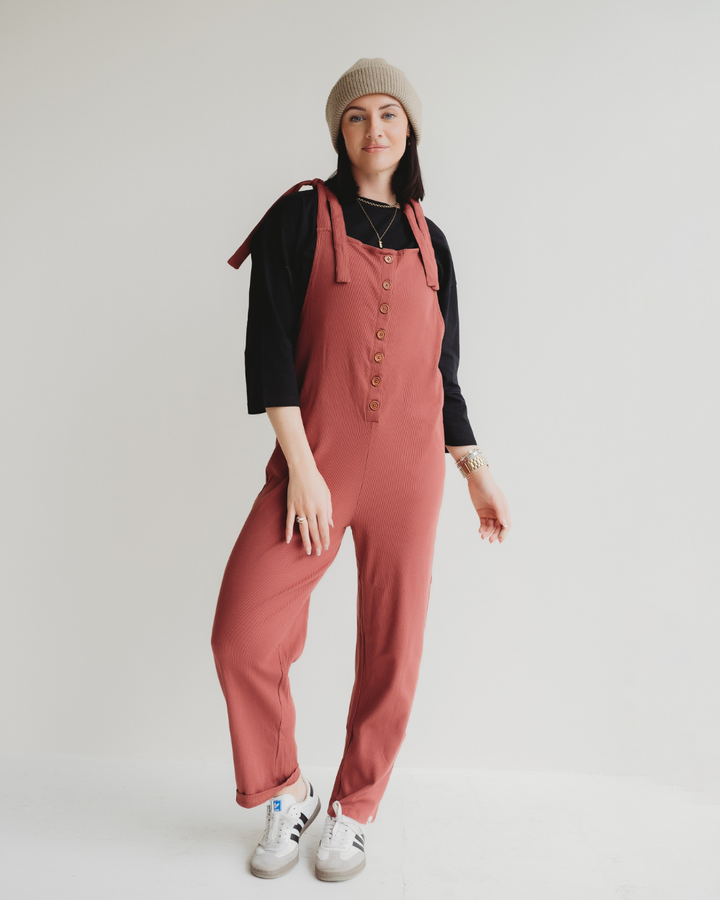 Wearing a beige beanie, black long-sleeve shirt, and Erin Brick Dungarees made from sustainable organic cotton, the person stands against a plain background. They complete their relaxed look with white sneakers and a slight smile.