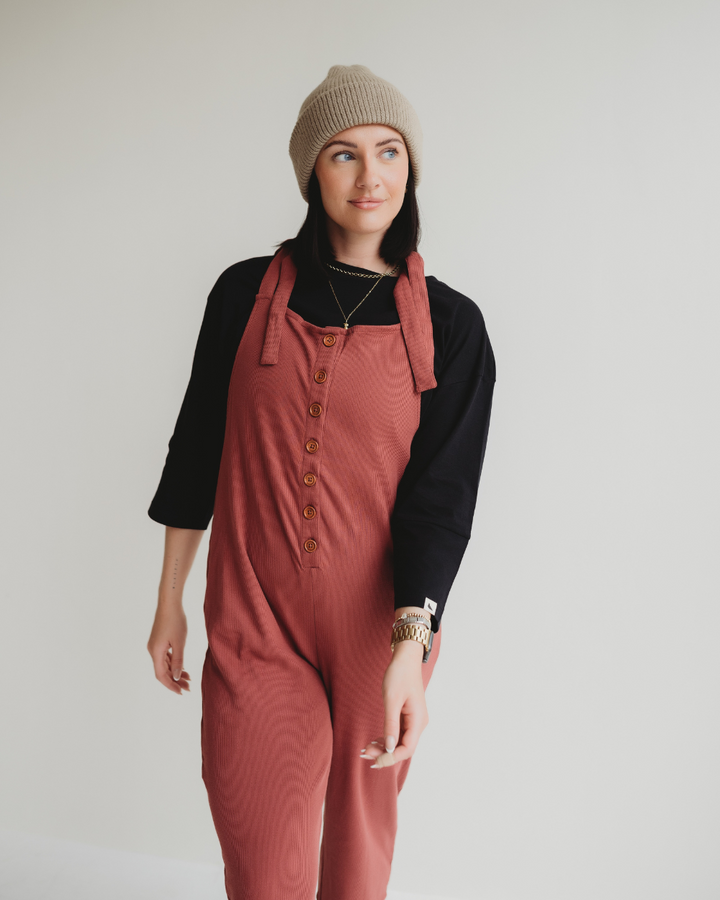 Wearing a beige beanie and black long-sleeve shirt, they don the Erin Brick Dungarees—a red ladies piece with buttons. With a slight smile, they stand against a plain white background, embodying effortless style and comfort.