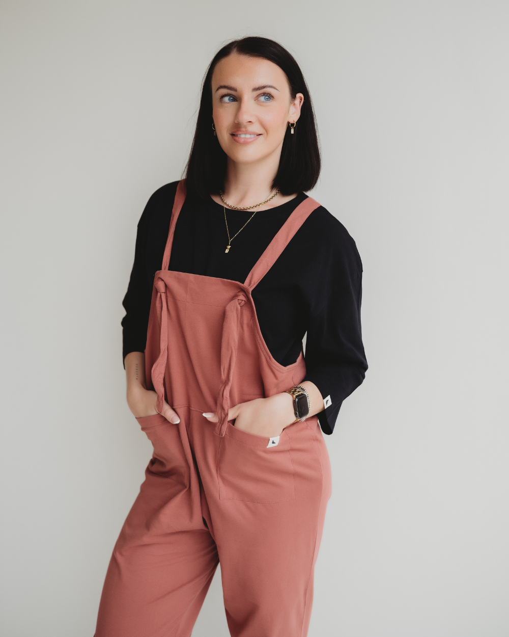 A person with shoulder-length black hair smiles against a white background, wearing a black top and rose-colored Lila Brick Dungarees. They showcase their style as a conscious consumer, accessorized with a necklace and watch.