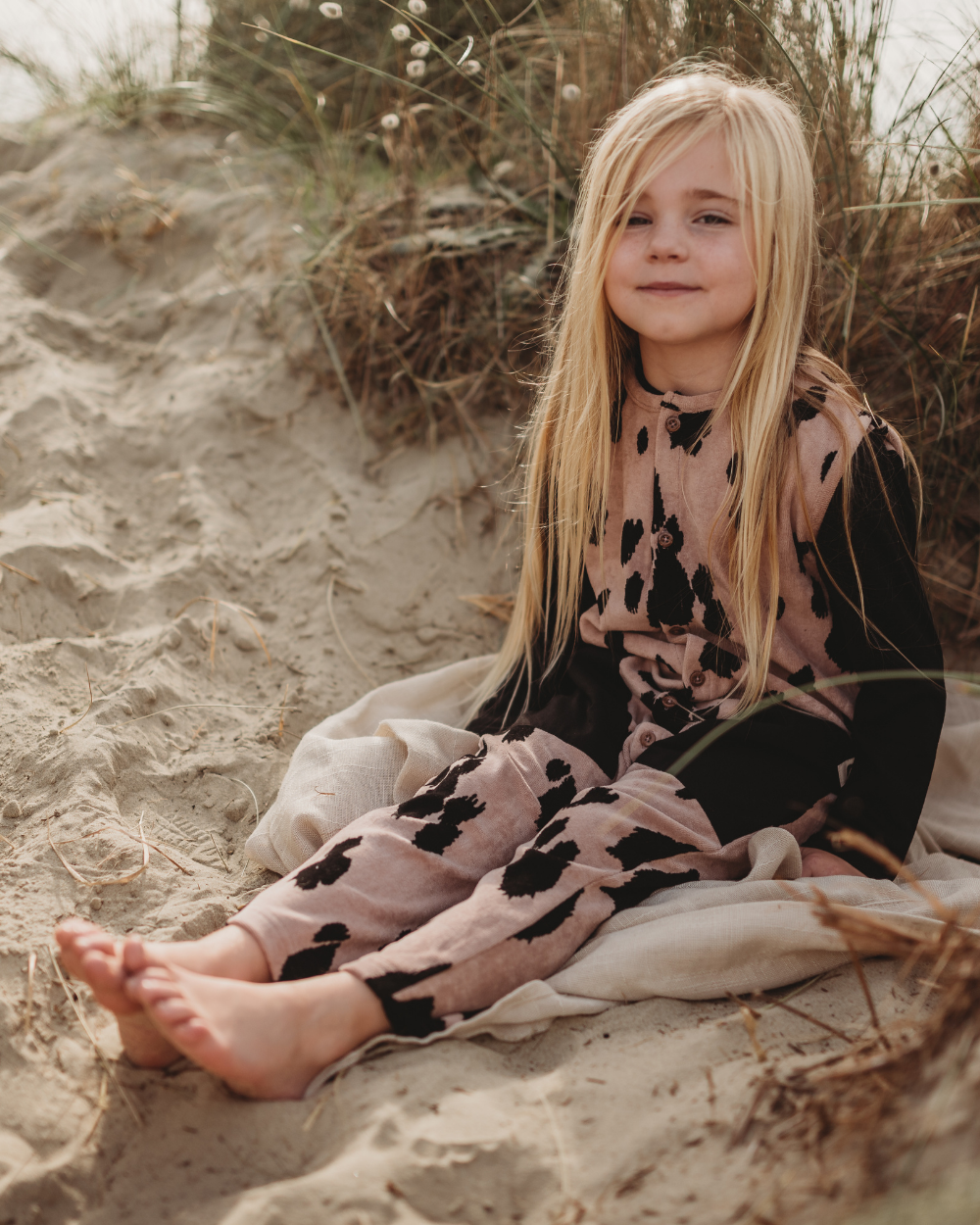 A young girl with long blonde hair sits on a blanket in the sand, wearing Pony Velour Dungarees crafted from organic fabric in earthy tones. She smiles softly at the camera as tall grasses sway behind her, creating a serene, beach-like scene.