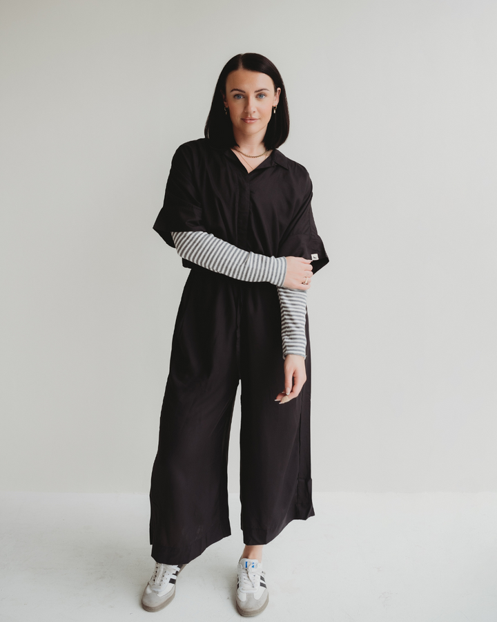 A person with shoulder-length dark hair stands against a white backdrop, wearing the Iris Black Jumpsuit made from EcoVero viscose. Its layered over a gray and white striped long-sleeve shirt and paired with white sneakers, as they gaze at the camera neutrally.