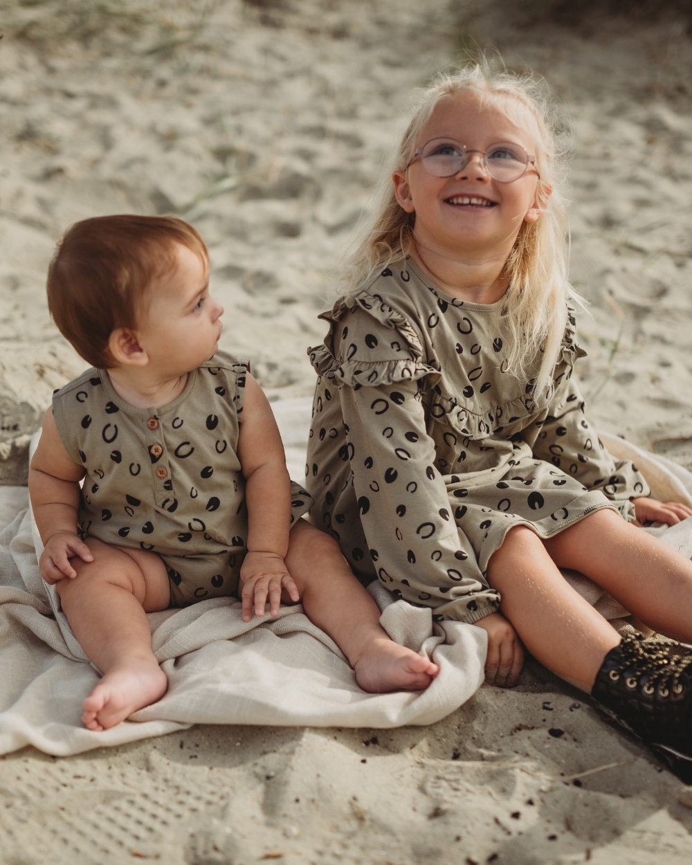 Two young children sit on a light blanket at the beach, both wearing beige Clip Clop Dresses featuring black prints. The right child, with glasses, smiles while looking up. Made from 100% GOTS organic cotton, the younger child glances at them.