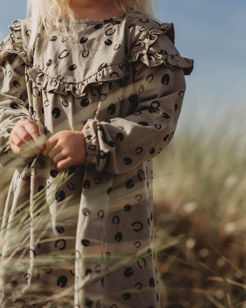 A child with long blond hair stands in tall grass, wearing a beige Clip Clop Dress made from 100% GOTS organic cotton. It features black polka dots and ruffled shoulders. Against a clear blue sky, the dress captures playful elegance.