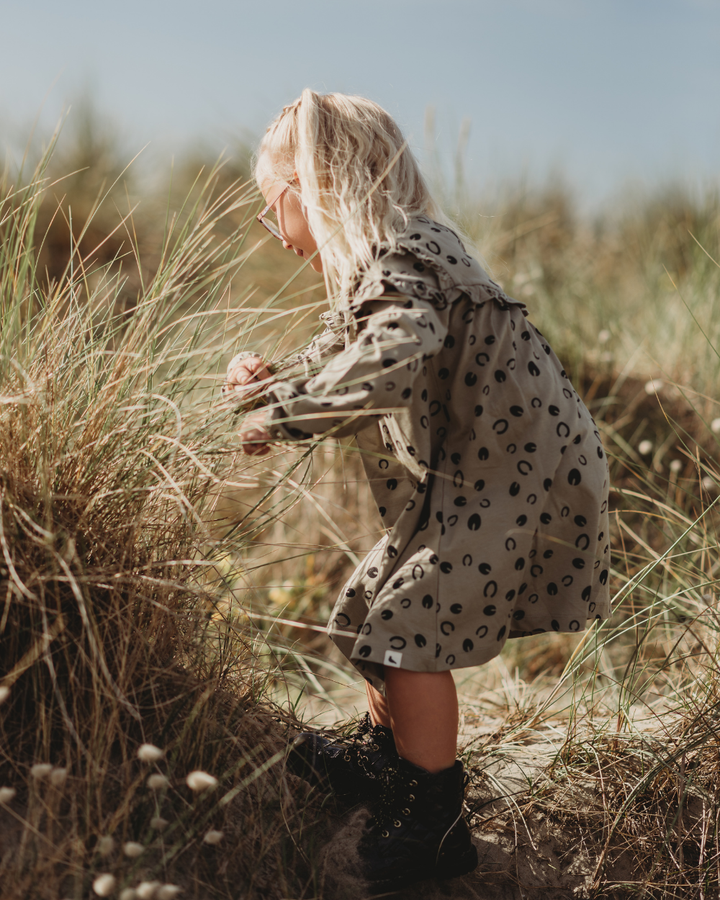 A young child with long blonde hair, wearing sunglasses and a Clip Clop Dress made of 100% GOTS organic cotton, explores the tall grass in a sandy dune. The clear blue sky suggests a sunny day.