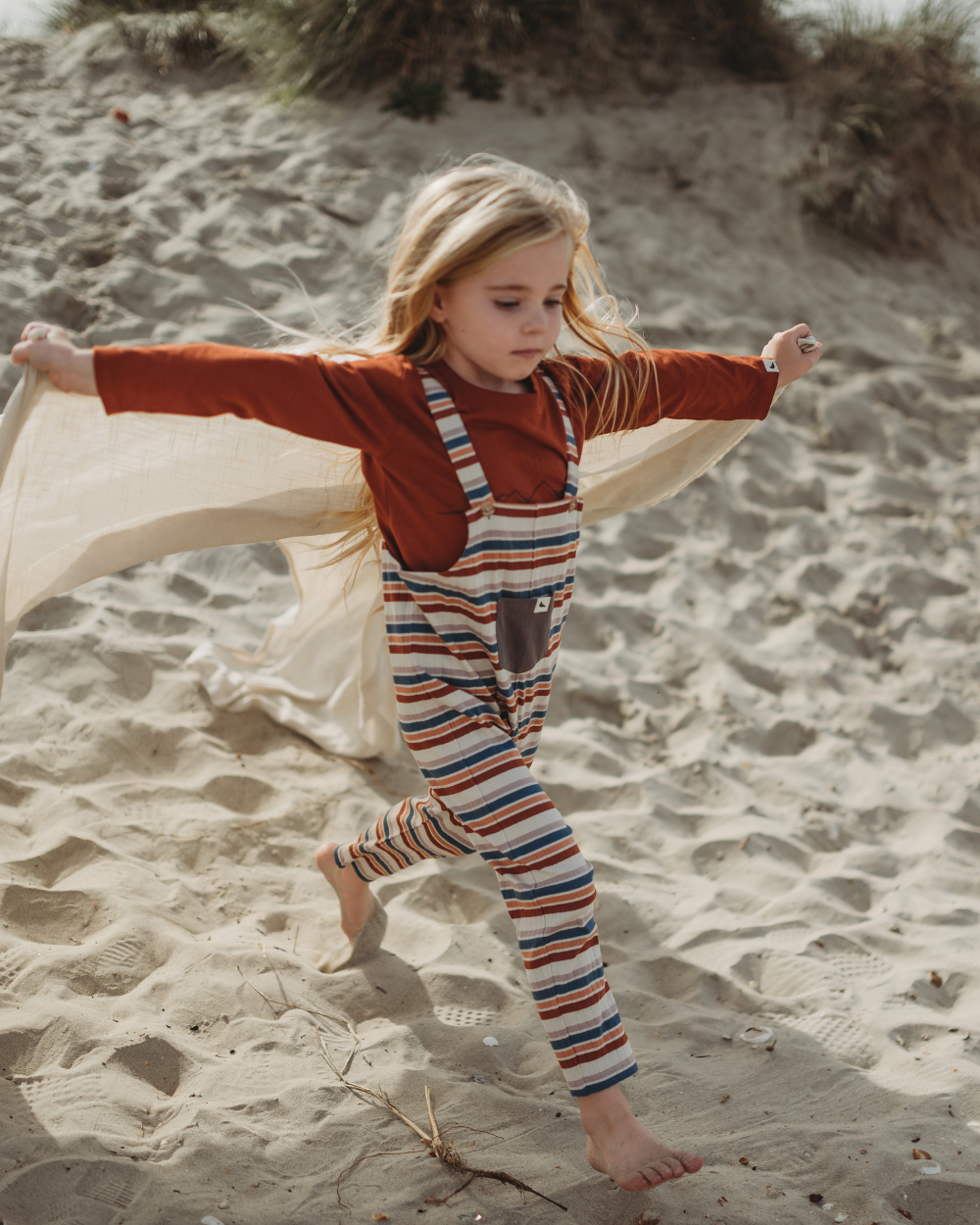 Barefoot on a sandy beach, a young girl with long hair runs joyfully. Her organic cotton Multi Stripe Dungarees flutter in the breeze as she holds a white cloth behind her, imagining flight. Dune grass sways in the background, symbolizing her commitment to sustainable, playful adventures.