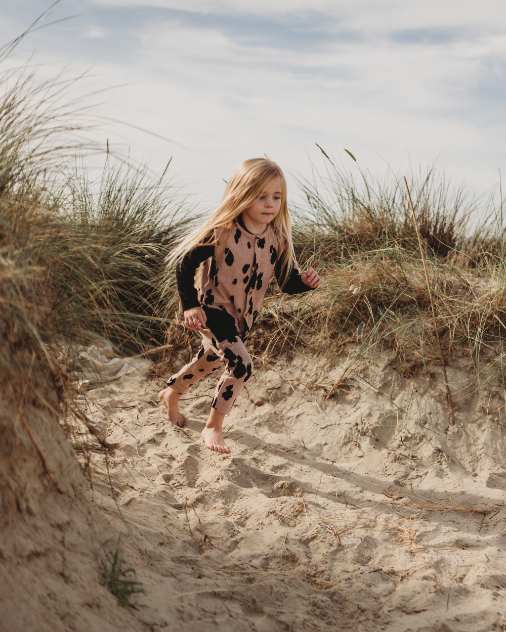 A young child with long blonde hair runs barefoot through sandy dunes in Pony Velour Dungarees. The black and brown patterned outfit complements the swaying tall grasses and cloudy sky, reflecting a sustainable wardrobe choice.