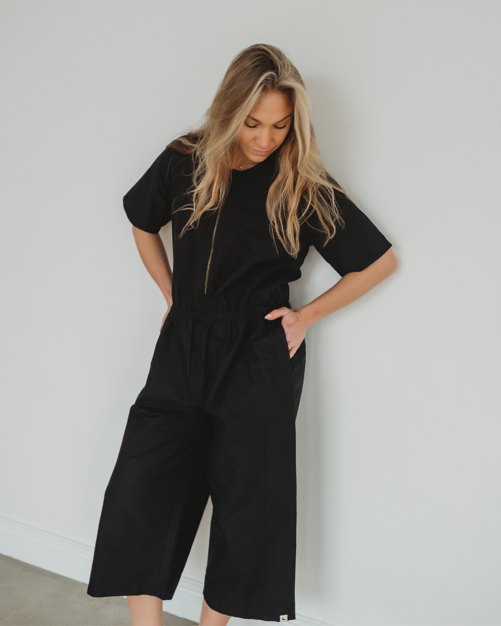 A person with long blonde hair, looking down with hands in pockets, stands against a white wall wearing the loose-fitting Ethel Twill Black Jumpsuit. Made from organic cotton, this short-sleeve outfit features sustainable summer style with wide-leg pants.