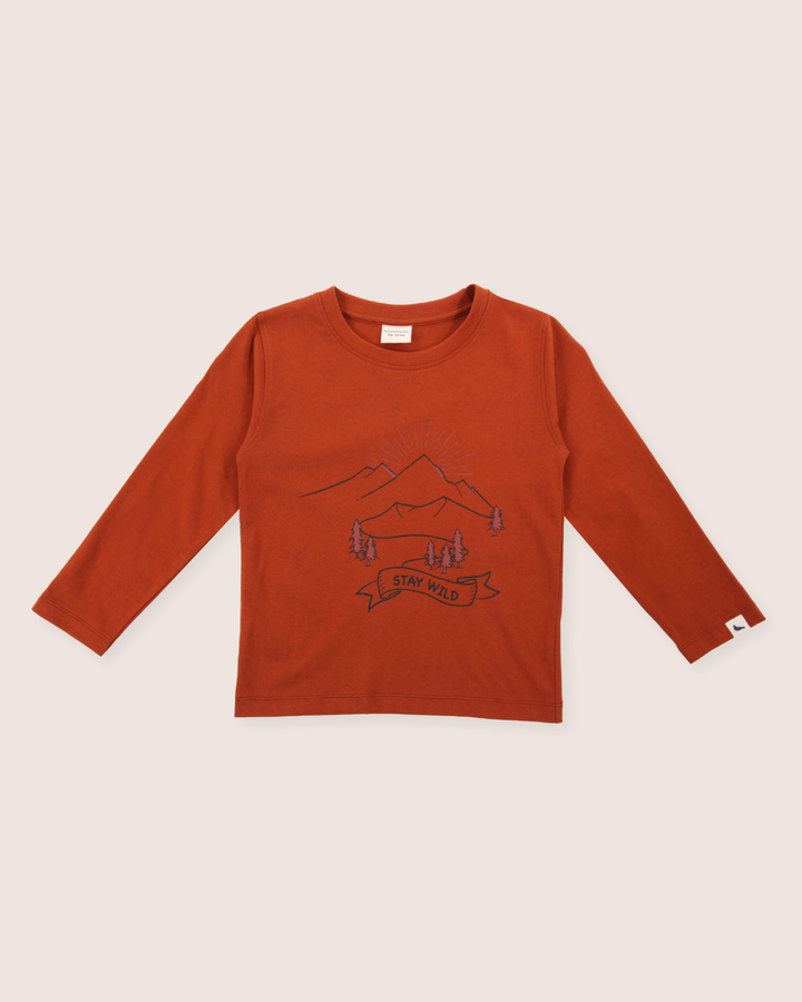 The Wild Mountains Top is a rust-colored, long-sleeved shirt made from organic cotton, featuring a mountain and tree design with the phrase Stay Wild in a banner below.