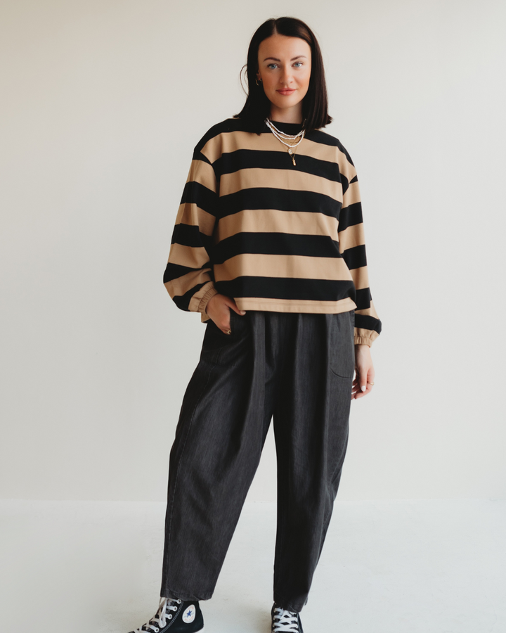 A person with shoulder-length dark hair confidently poses against a light background, wearing the Ivy Striped Sweatshirt in beige and black, paired with dark loose-fitting pants and black sneakers. Their hands are in their pockets, and they are smiling warmly.