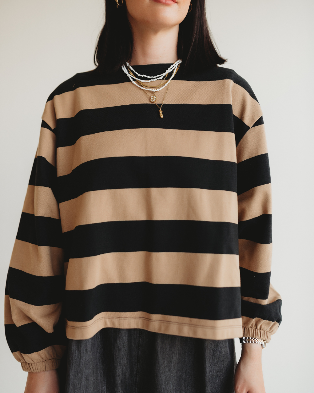 The woman dons an Ivy Striped Sweatshirt in black and tan, made of organic cotton, accessorized with layered necklaces. Her dark hair contrasts the white backdrop, highlighting her sustainable fashion sense.