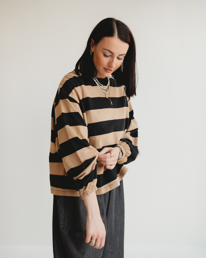 A woman with shoulder-length dark hair stands against a plain background, wearing the Ivy Striped Sweatshirt in beige and black made of organic cotton and dark pants. She appears to adjust her balloon sleeve while accessorizing with layered necklaces and hoop earrings.