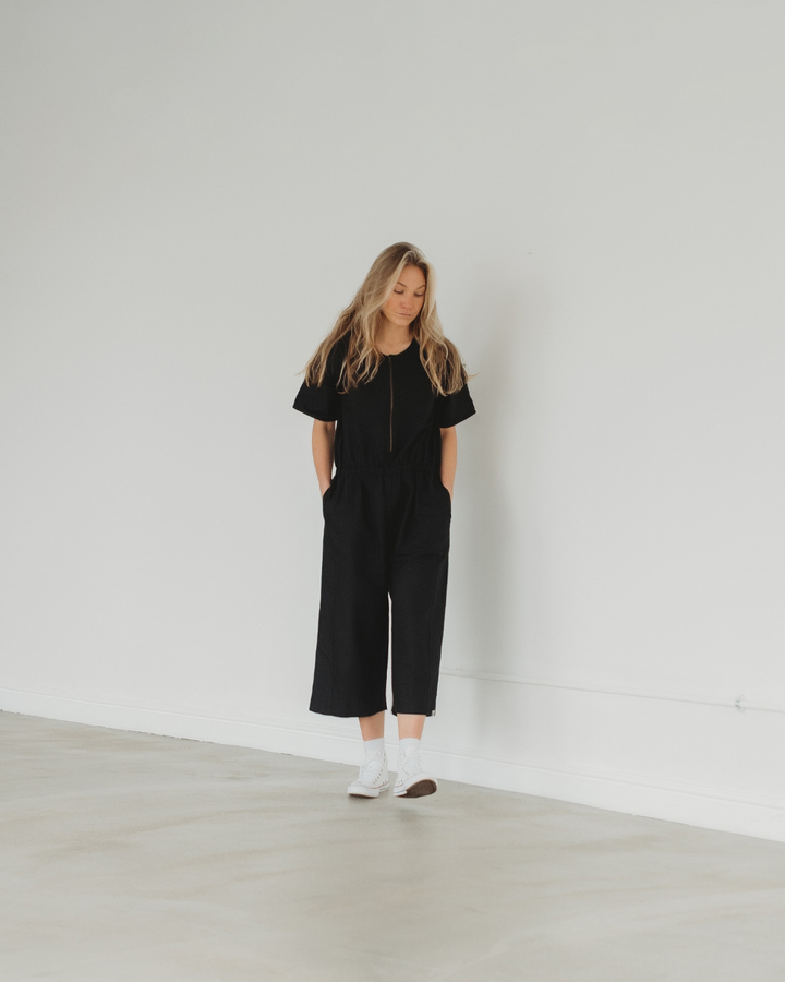 In a minimalist room, a person with long blonde hair embodies sustainable summer style, wearing an Ethel Twill Black Jumpsuit and white sneakers. They lean against a white wall in a relaxed posture.