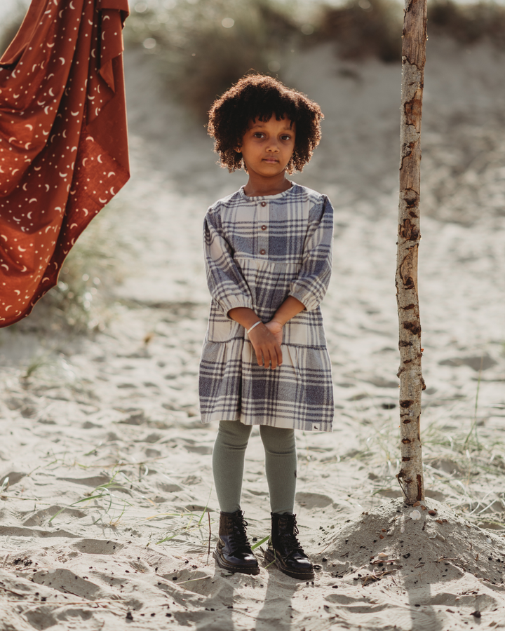 A young girl stands on a sandy path, wearing a Brushed Check Dress with green leggings and black boots. Nearby, patterned brown fabric hangs over a branch. The blurred green background creates a picturesque scene perfect for capsule wardrobes.