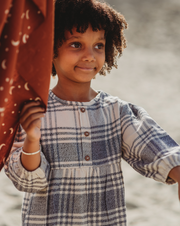 A smiling child with curly hair holds a rust-colored curtain adorned with moons and stars. They stand in a sunlit outdoor setting wearing an organic cotton Brushed Check Dress, encapsulating the essence of capsule wardrobes.