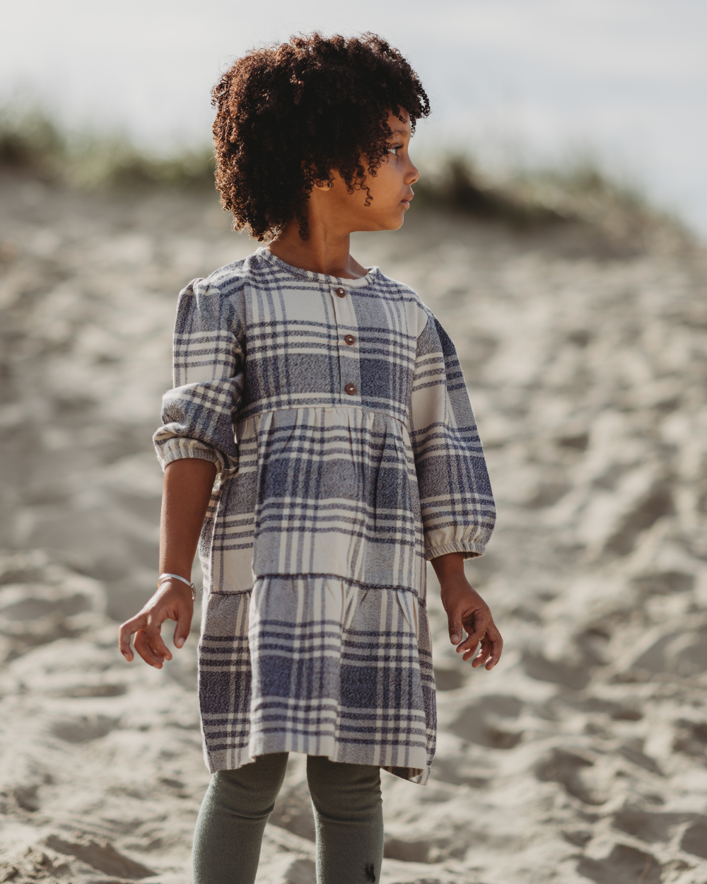 A young girl on a beach looks right, her curly hair flowing. She wears a Brushed Check Dress with long sleeves, organic cotton material, and a tiered skirt paired with dark leggings. Sand dunes and grass form the blurred backdrop.