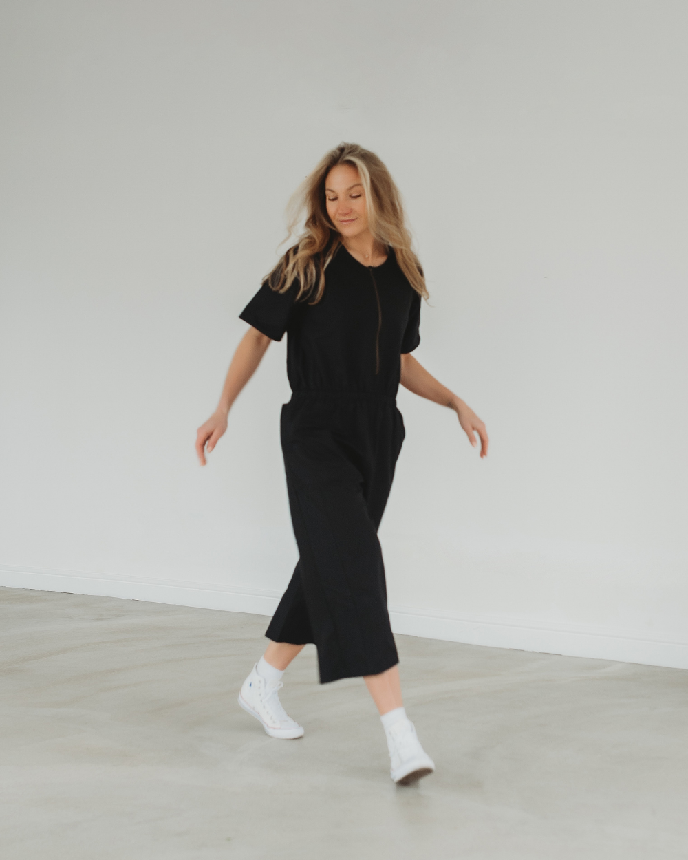 A person with long hair is mid-stride against a plain, light background, wearing the Ethel Twill Black Jumpsuit and white sneakers, embodying sustainable summer style.