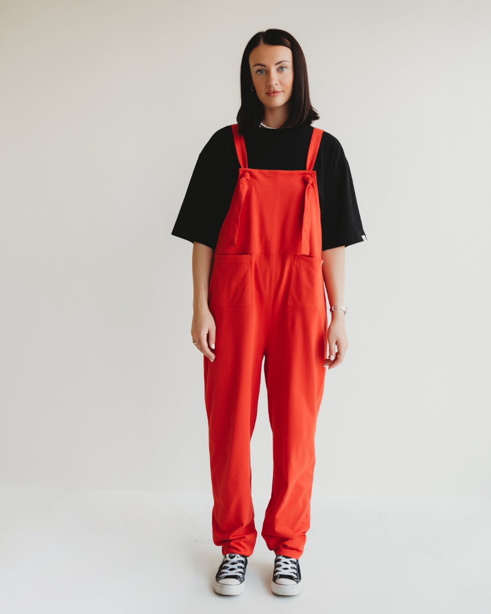 Standing against a plain white background, a person dons Lila Plain Red Dungarees made of organic cotton and a black t-shirt. With black and white sneakers completing the look, they exemplify conscious consumerism while gazing neutrally at the camera.