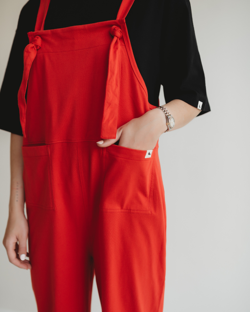 A stylish individual exudes eco-friendly fashion in bright red Lila Plain Red Dungarees with hands in the pockets, paired with a black shirt. A silver watch gleams on their wrist against a white backdrop, ideal for the conscious consumer.