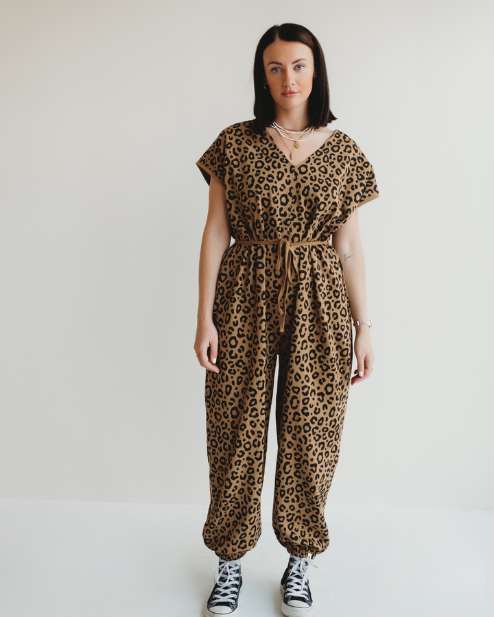 A person in a Rita Tie Back Mini Animal Print Jumpsuit with elastic hems stands against a white background. They have long dark hair, wear black and white sneakers, their hands are at their sides, and they gaze directly at the camera.