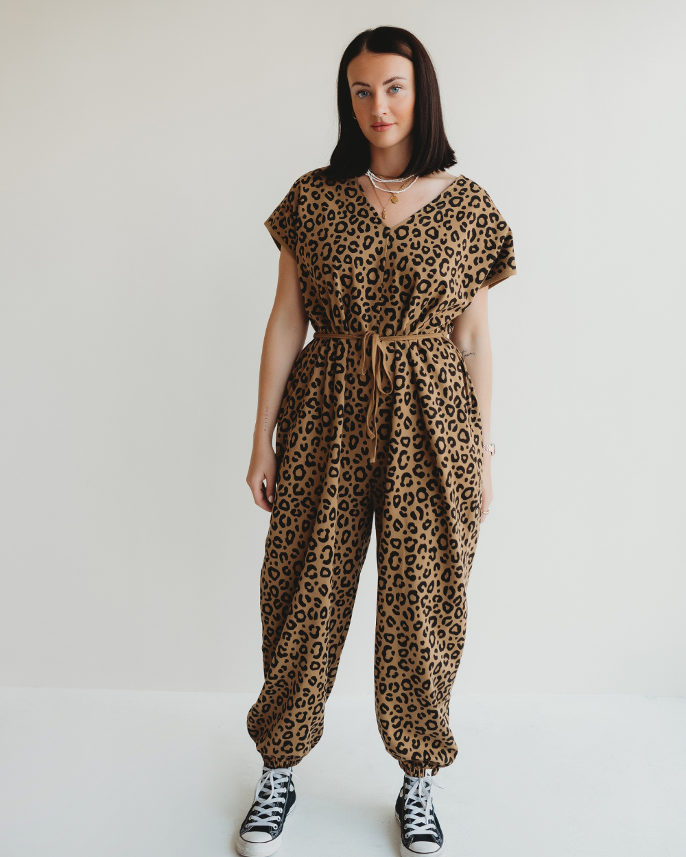 Against a white background, a person with long dark hair adopts a relaxed posture, gazing at the camera. They are wearing the Rita Tie Back Mini Animal Print Jumpsuit made of organic cotton paired with black sneakers.