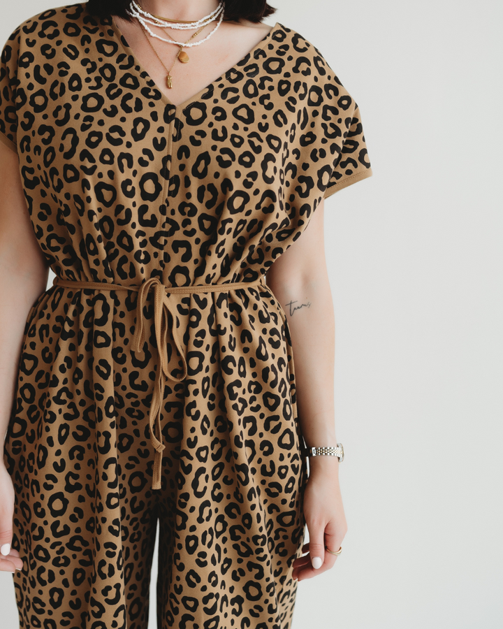 A person wears the Rita Tie Back Mini Animal Print Jumpsuit in brown with short sleeves and a tied waist, crafted from organic cotton. They accessorize with layered necklaces and a wristwatch, standing against a plain white background. Their face is not visible.