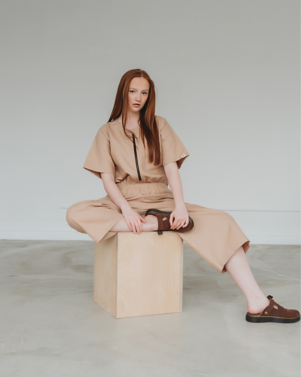 A person with long red hair sits casually on a wooden cube against a plain background, wearing the Ethel Plain Clay Jumpsuit. This tan organic cotton outfit offers a zip-up-and-go style, perfectly paired with brown clogs for effortless flair.