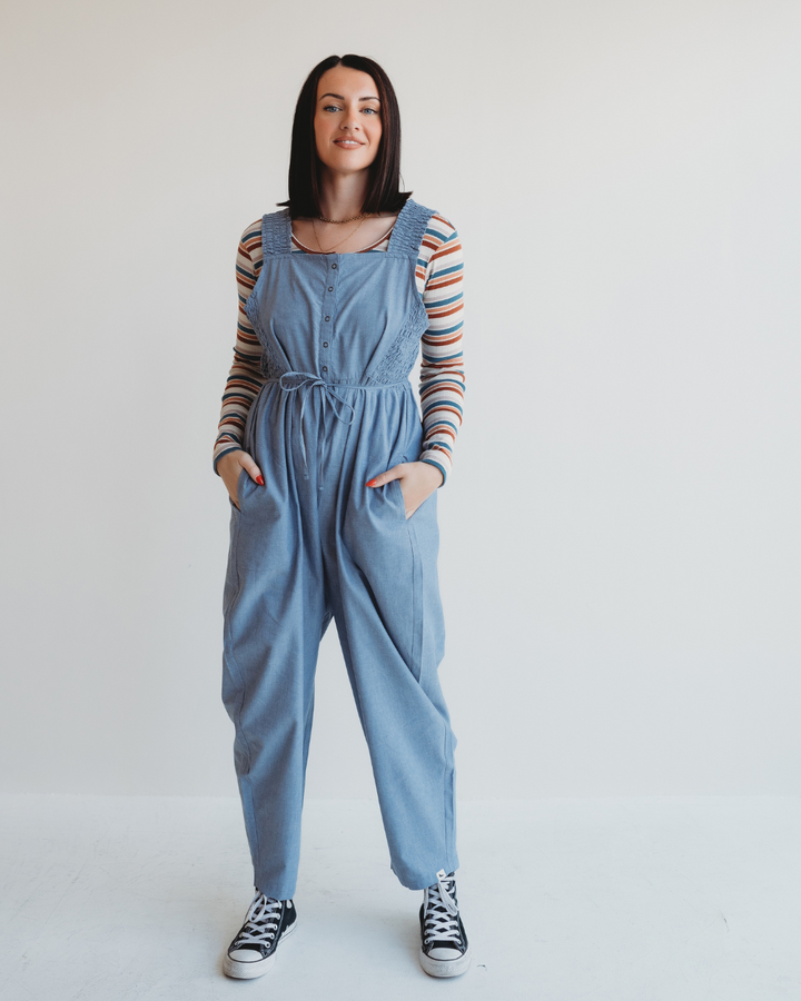 A person with long dark hair stands against a plain background, wearing Alice Chambray Dungarees over a striped long-sleeve shirt and black high-top sneakers. They have their hands in the dungaree pockets and are smiling.