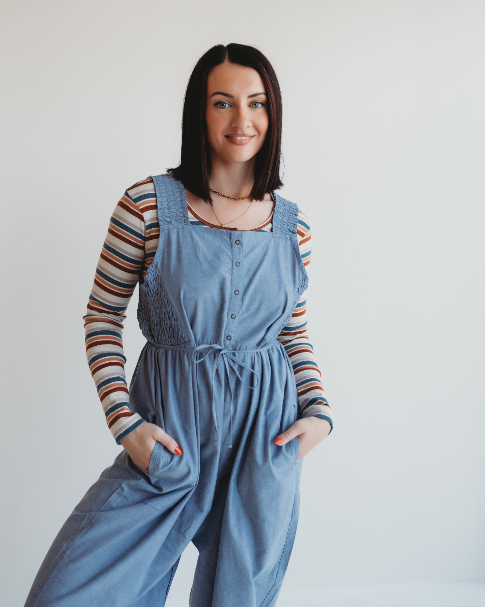 A woman with straight black hair smiles against a plain white background, wearing an Alice Chambray Dungarees over a striped long-sleeve top, hands in pockets and front buttons visible.