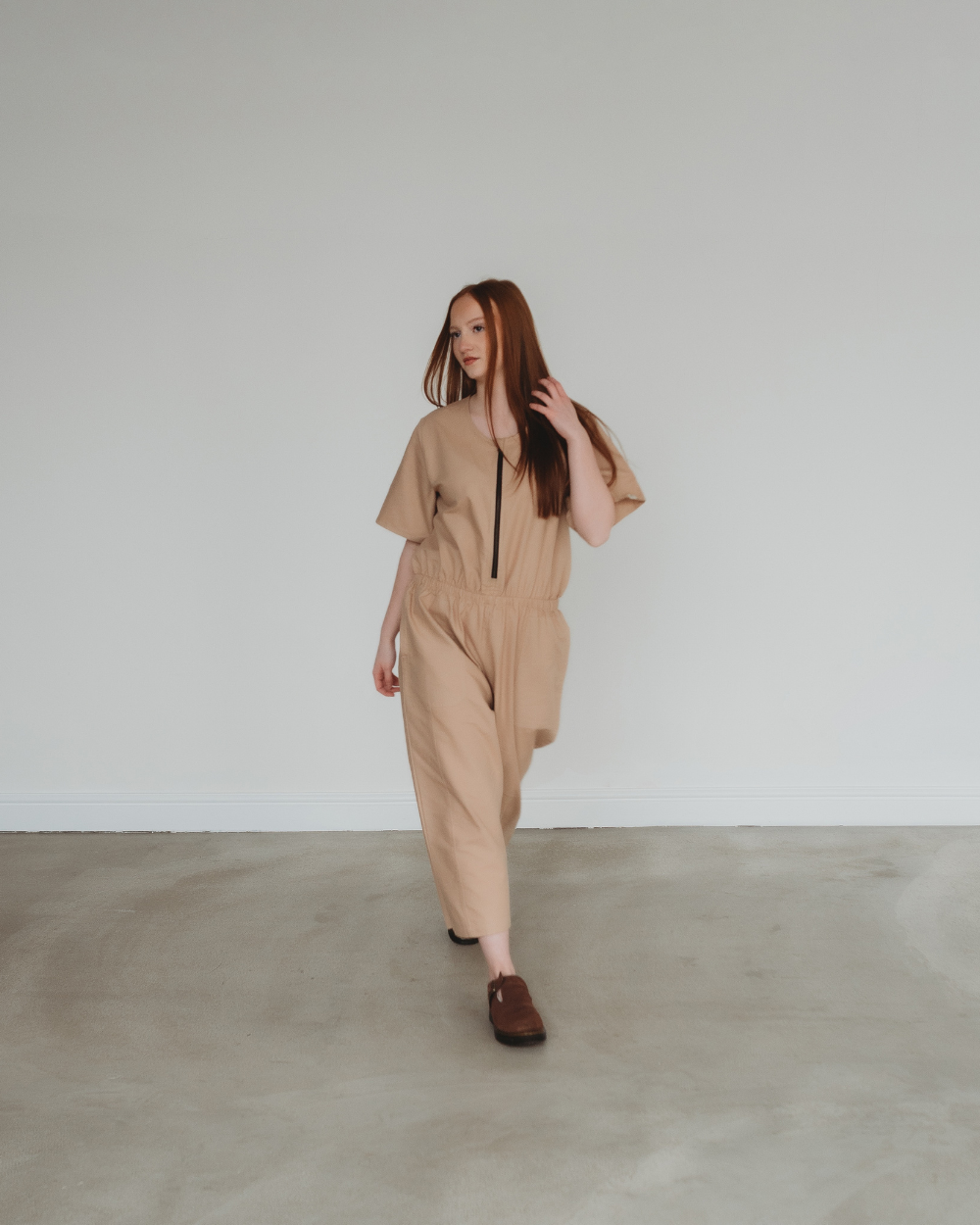 In a loose-fitting Ethel Plain Clay Jumpsuit, a person with long hair exudes confidence as they walk on a smooth, light gray floor against a white wall. They complete their effortlessly chic look with dark brown shoes.