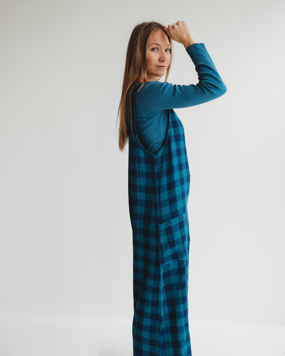 Wearing a blue long-sleeve shirt paired with Grace Check & Stripe Reversible Navy Jumpsuit, an individual with long hair stands against a plain white background, looking over their shoulder gently. This stylish outfit seamlessly combines eco-consciousness for a perfect sustainable wardrobe.