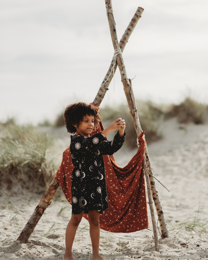 A child stands barefoot on a sandy beach, wearing the Astrology Dress, featuring sun and moon patterns. They play with a wooden teepee covered in organic cord fabric, as grassy dunes rise against a cloudy sky in the background.