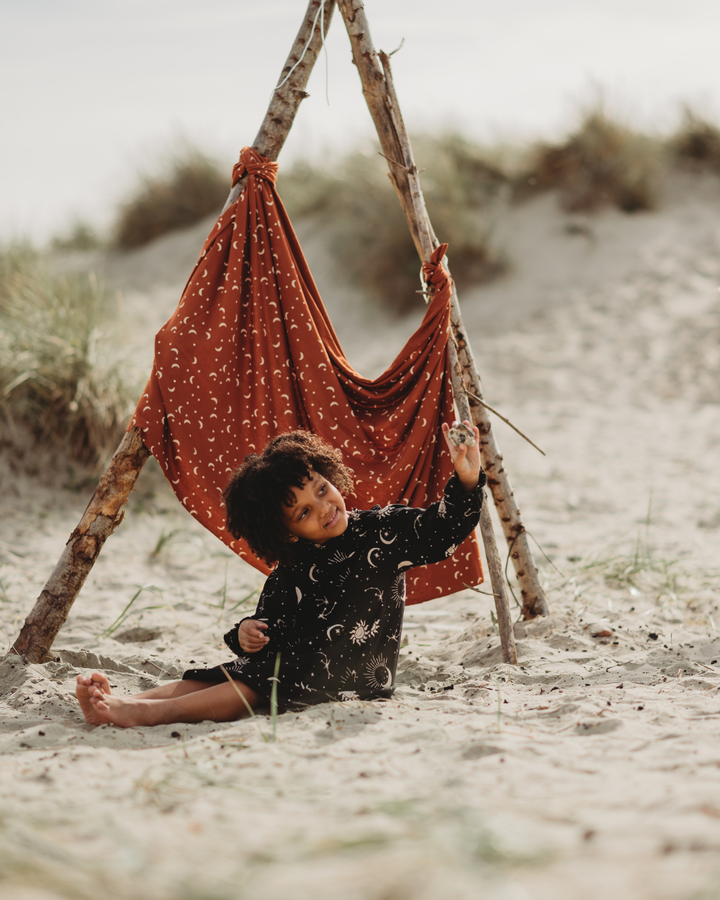 A child with curly hair sits on sandy ground by a stick frame tent, dressed in the charming Astrology Dress made from organic cord fabric. The child holds a small object, smiling with grassy dunes blurred in the background.