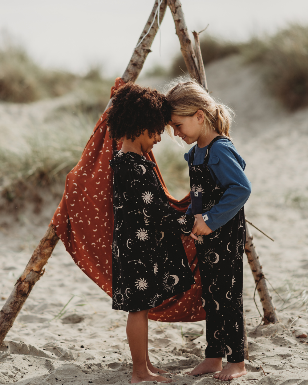 Two children stand barefoot on a sandy beach, touching foreheads and holding hands in Astrology Dresses. Behind them is a makeshift teepee crafted from branches, draped with an organic cord fabric adorned with a stars-and-moons astrology print.