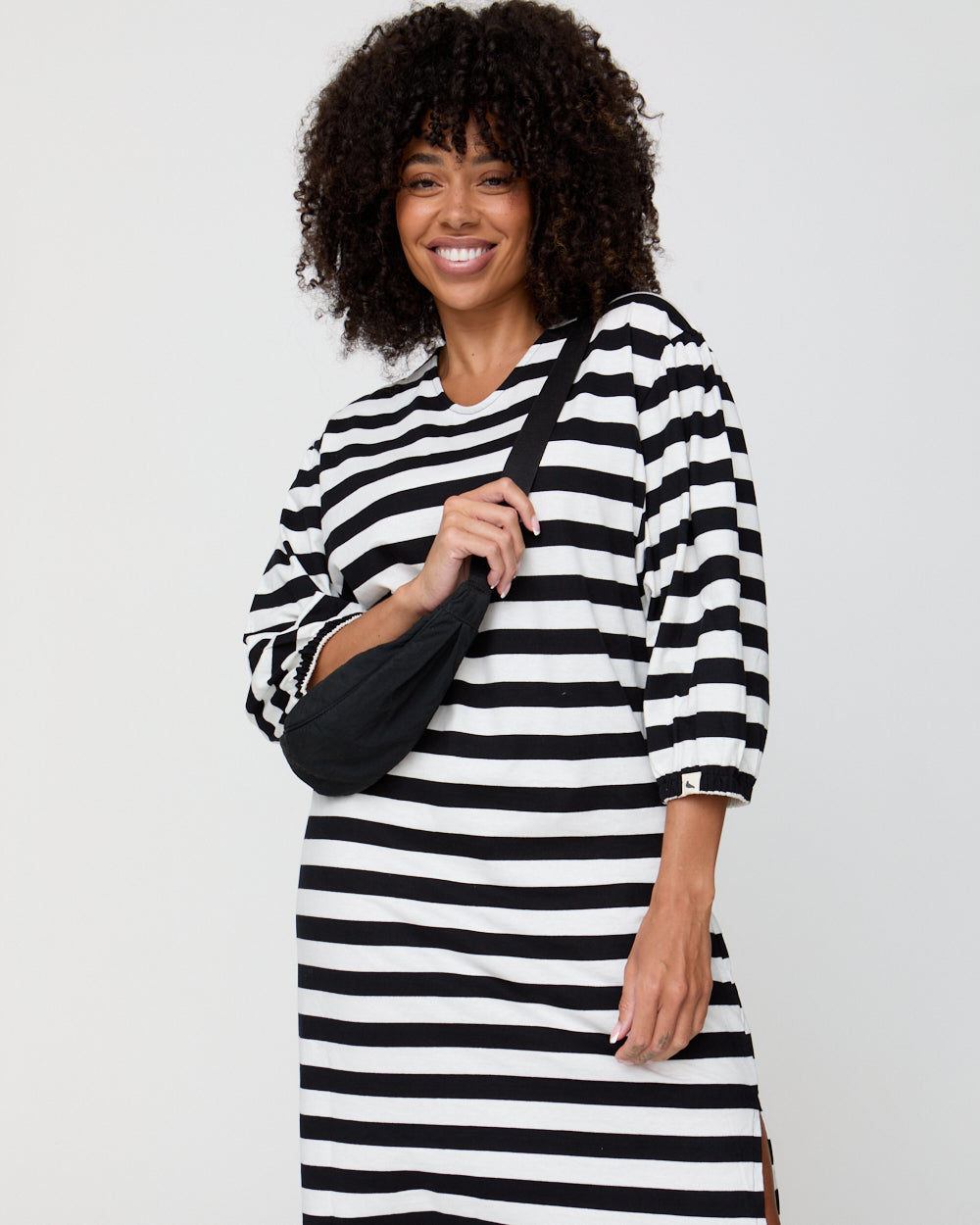 Woman wearing a black and white striped dress holding a black bag against a plain background
