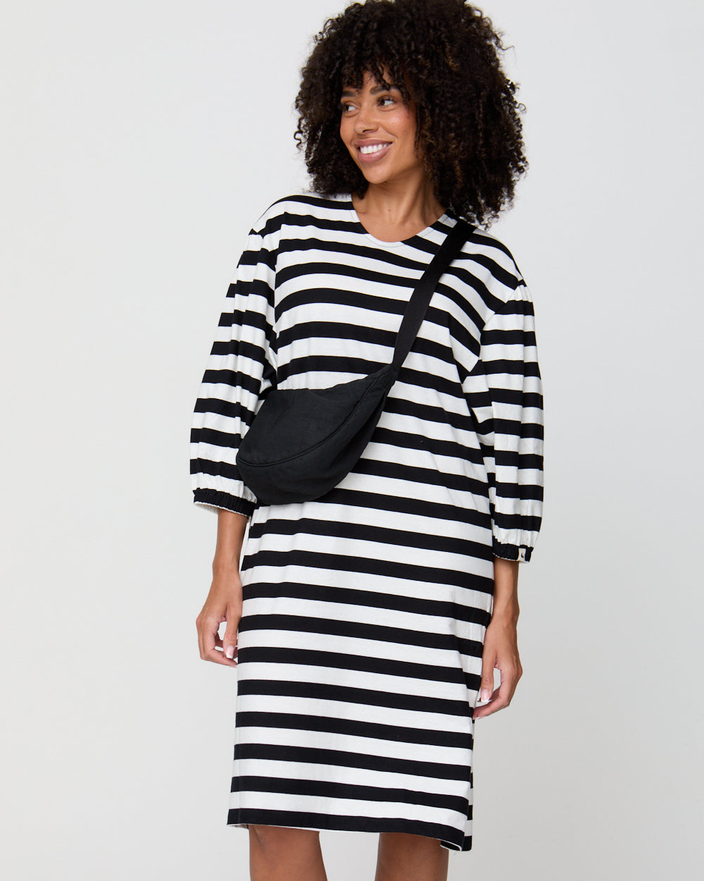 Woman wearing a black and white striped dress with a black crossbody bag on a plain background