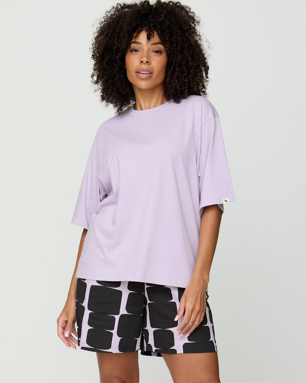 Woman wearing a light purple oversized t-shirt and black patterned shorts on a white background