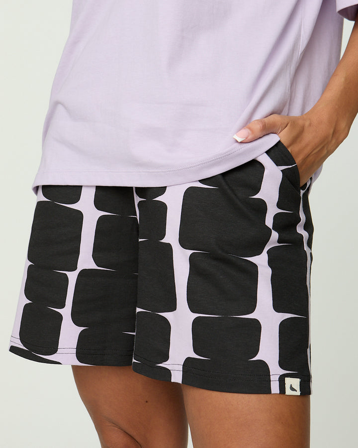Person wearing black and white patterned shorts with a light purple top on a plain background