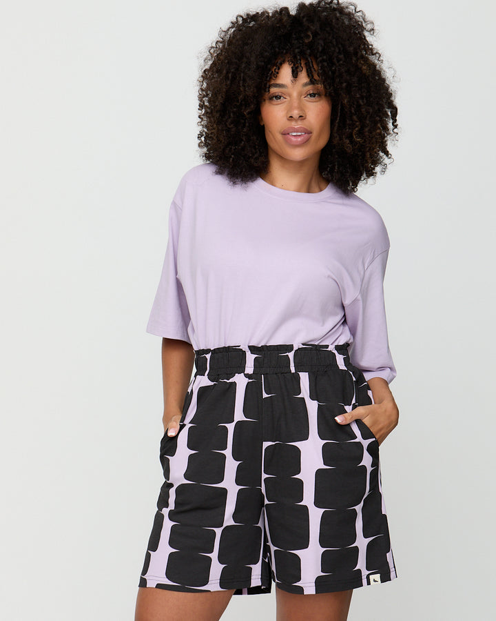 Woman wearing a light purple top and black and white patterned shorts on a plain background