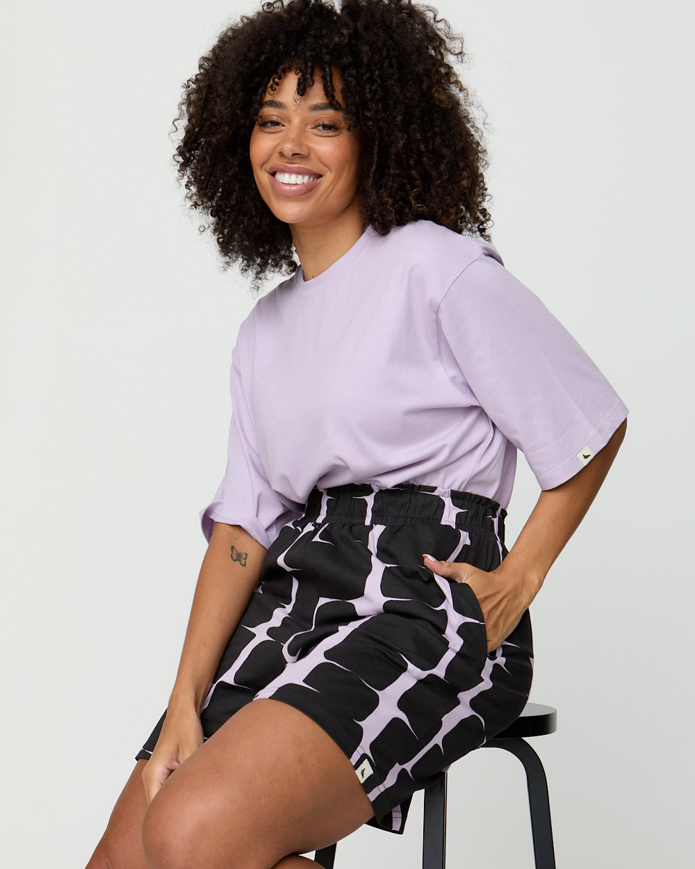 Woman wearing a light purple t-shirt and black patterned skirt sitting on a stool against a plain background
