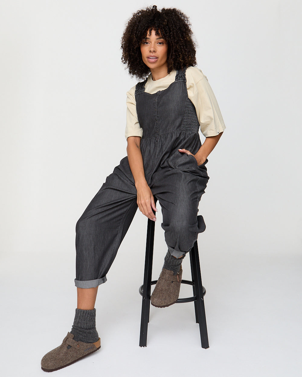 Person wearing dark gray overalls sitting on a black stool against a white background