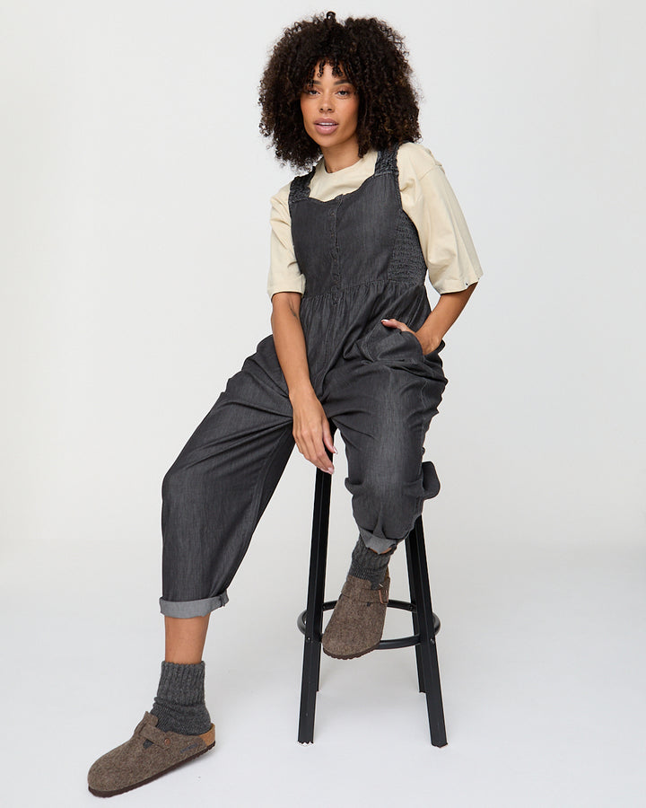 Person wearing dark gray overalls sitting on a black stool against a white background
