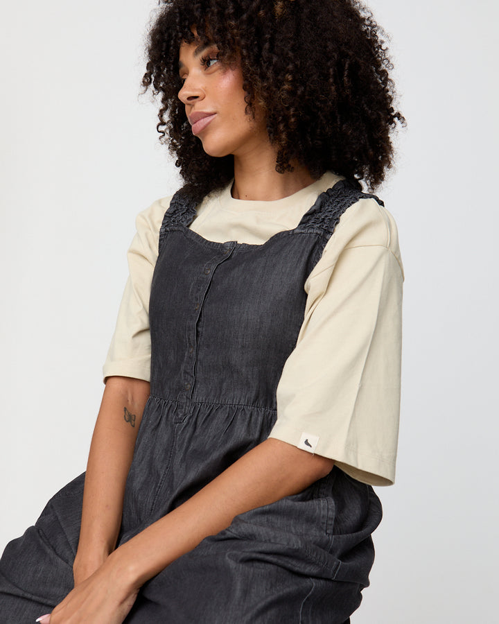 Woman wearing a black pinafore dress over a beige shirt on a plain background