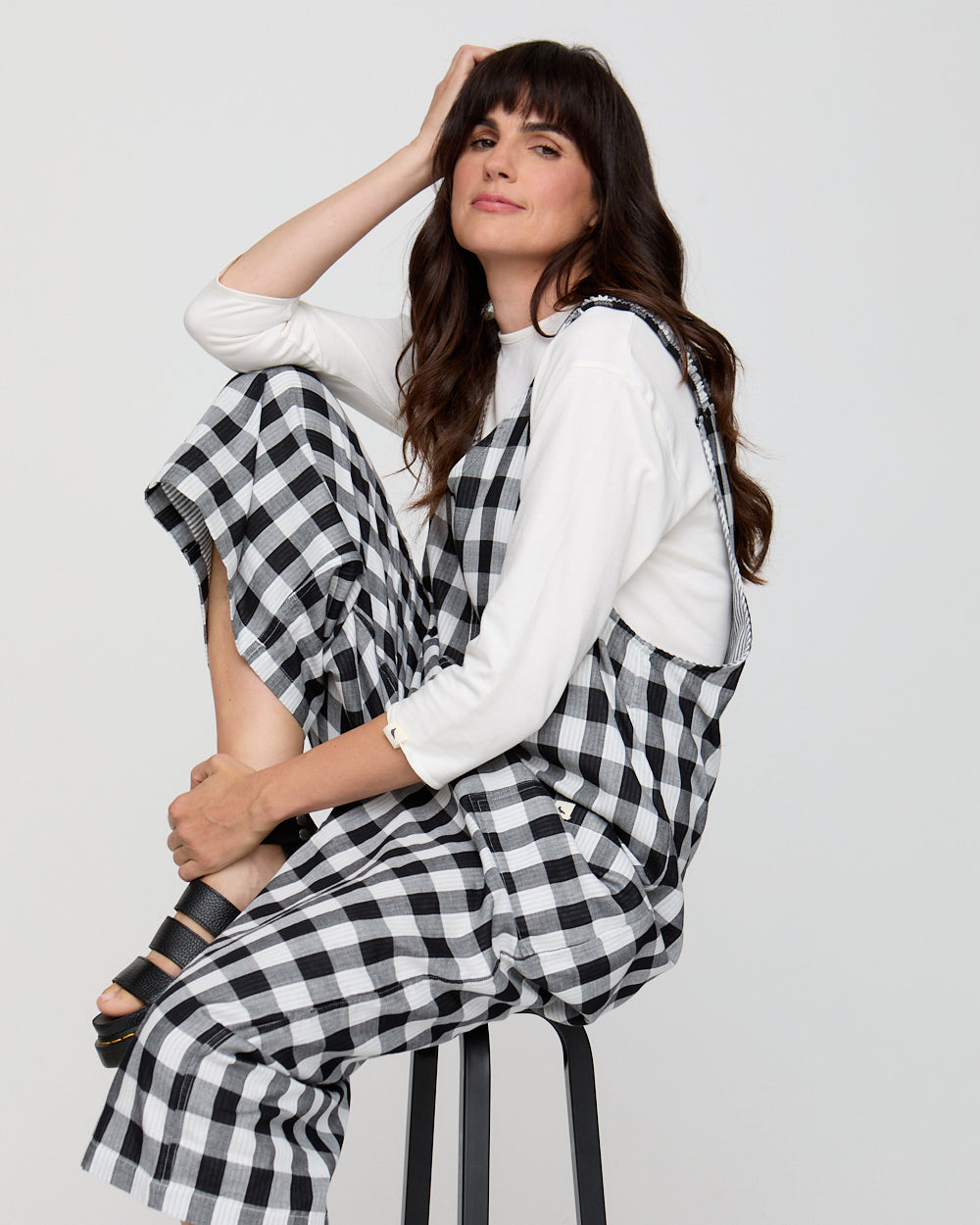 Woman wearing a white top and black and white checkered pants sitting on a stool against a plain background