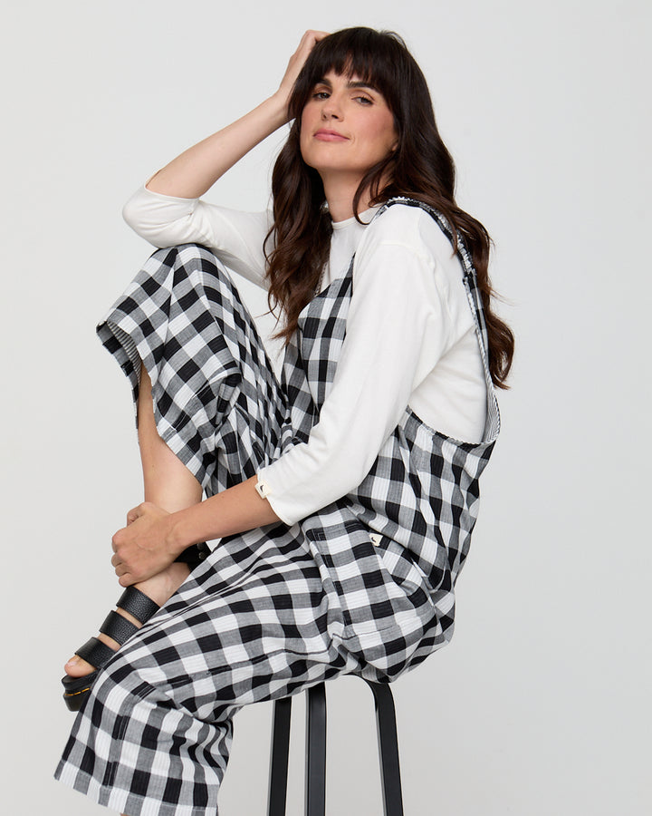 Woman wearing a white top and black and white checkered pants sitting on a stool against a plain background