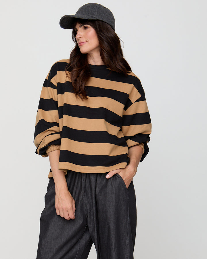 Woman wearing a black and beige striped sweater with dark pants against a plain background